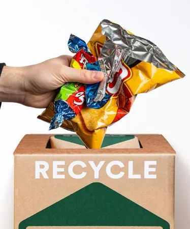 A hand dropping empty chip bags and candy wrappers into a TerraCycle Zero Waste Box marked Recycle